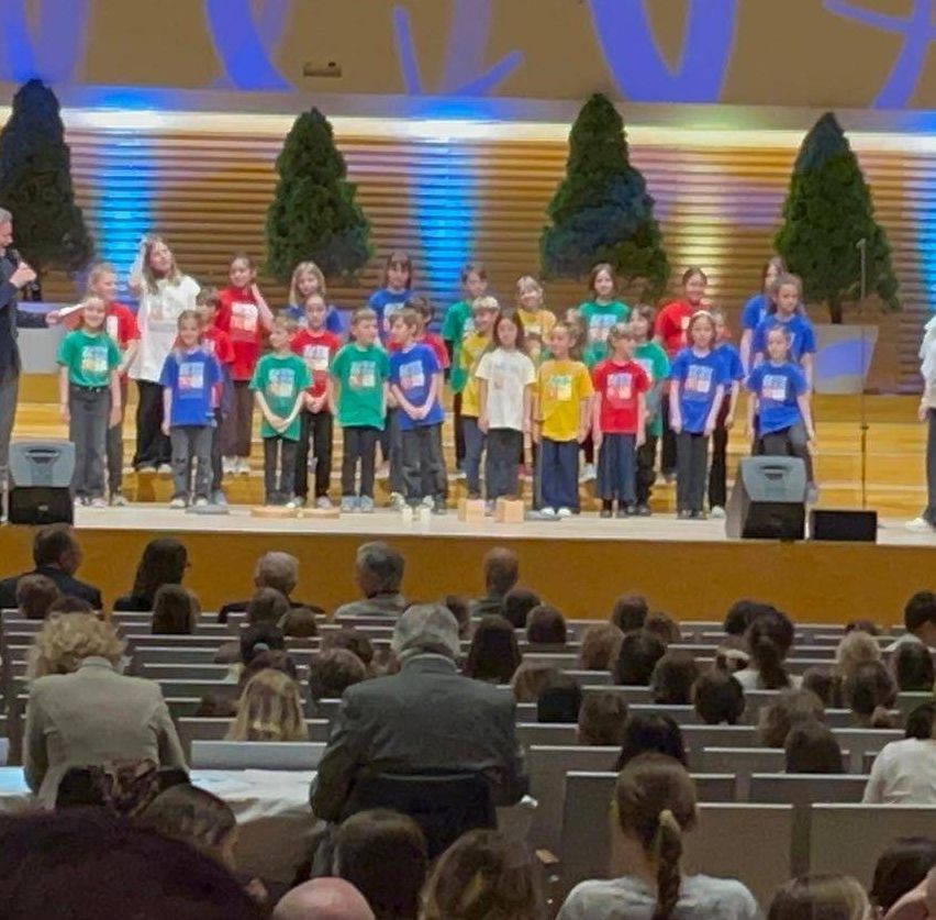 Ein Kinderchor in grünen, gelben und roten T-Shirts steht auf der Bühne. Eine Frau mit einem Mikrofon begleitet sie. Das Publikum sitzt in Reihen darunter. Bäume und blaue Beleuchtung im Hintergrund.