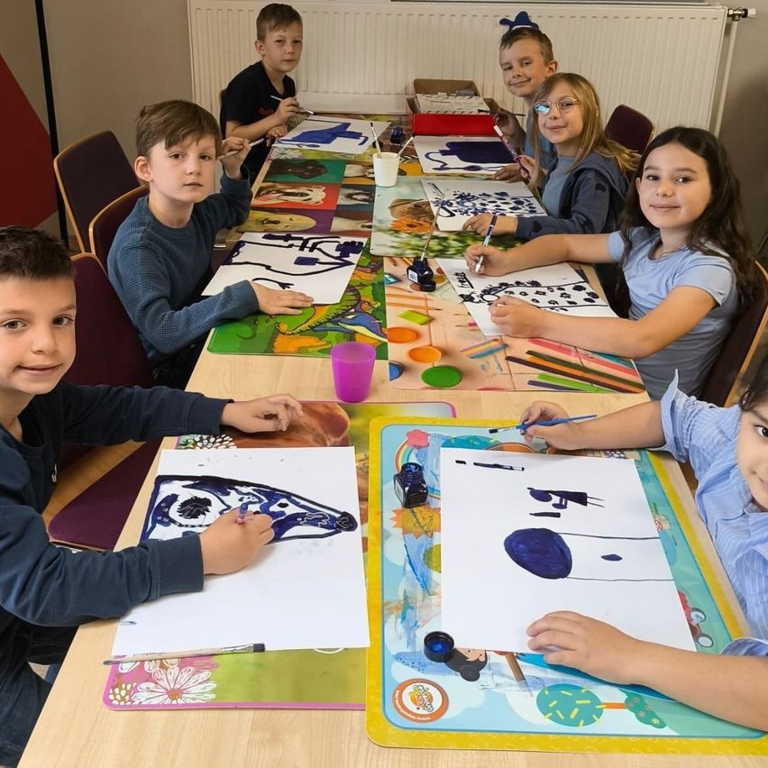 Eine Gruppe von Kindern sitzt um einen Tisch mit Malutensilien. Sie konzentrieren sich alle auf ihre eigenen Kunstwerke. Sie tragen alle Freizeitkleidung.