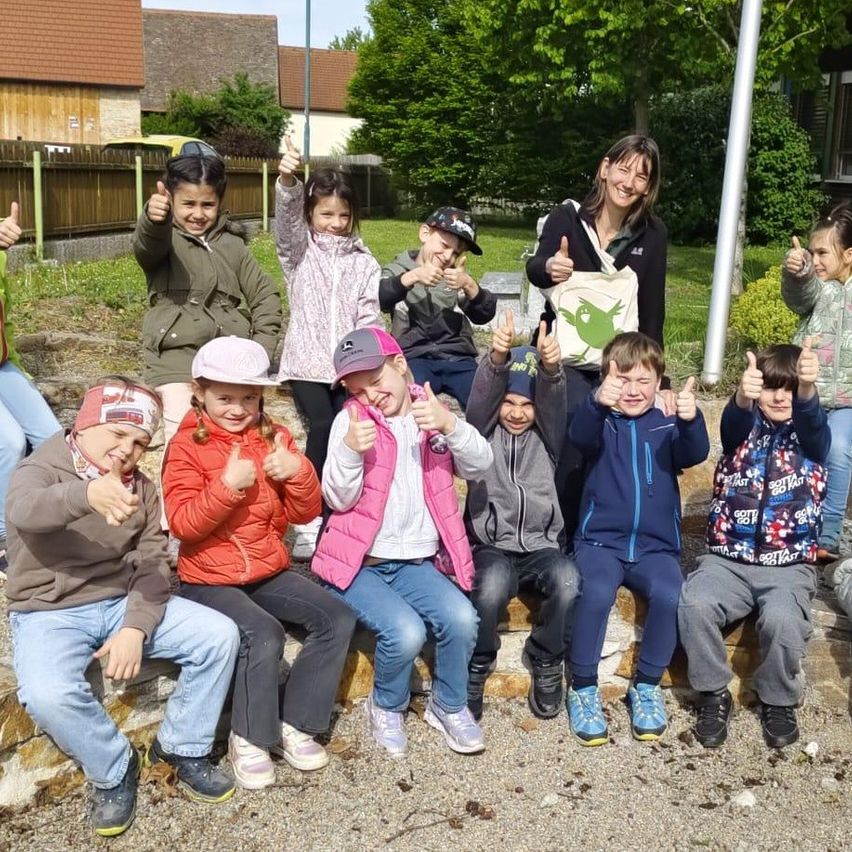 Gruppe von Kindern und Erwachsenen posiert für ein Foto im Freien. Sie sitzen auf einer Steinmauer, eine Frau steht hinter ihnen und hält eine Tasche. Die Kinder lächeln und zeigen Daumen hoch.