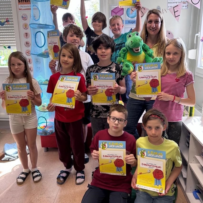 Eine Gruppe von Kindern und zwei Erwachsenen in einem Klassenzimmer. Sie halten Urkunden hoch und einer hat ein Krokodil-Plüschtier. Die Kinder tragen Freizeitkleidung.