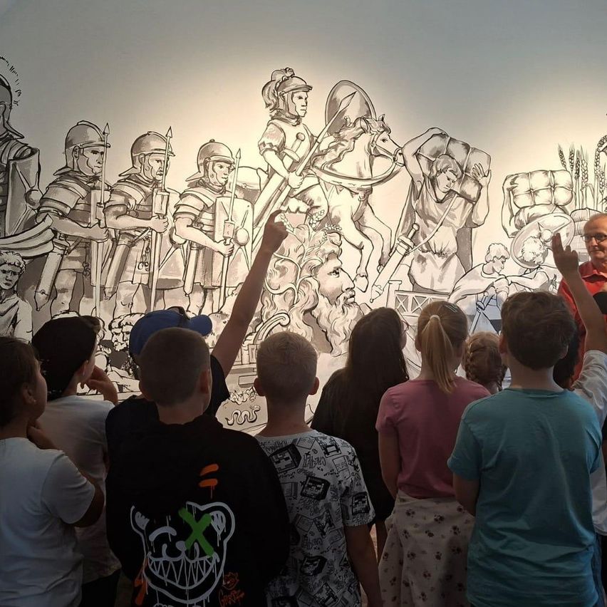 Eine Gruppe von Kindern und Erwachsenen versammelt sich um ein großes Wandgemälde, das Soldaten in historischer Kleidung darstellt, wobei eine Person auf das Bild zeigt.