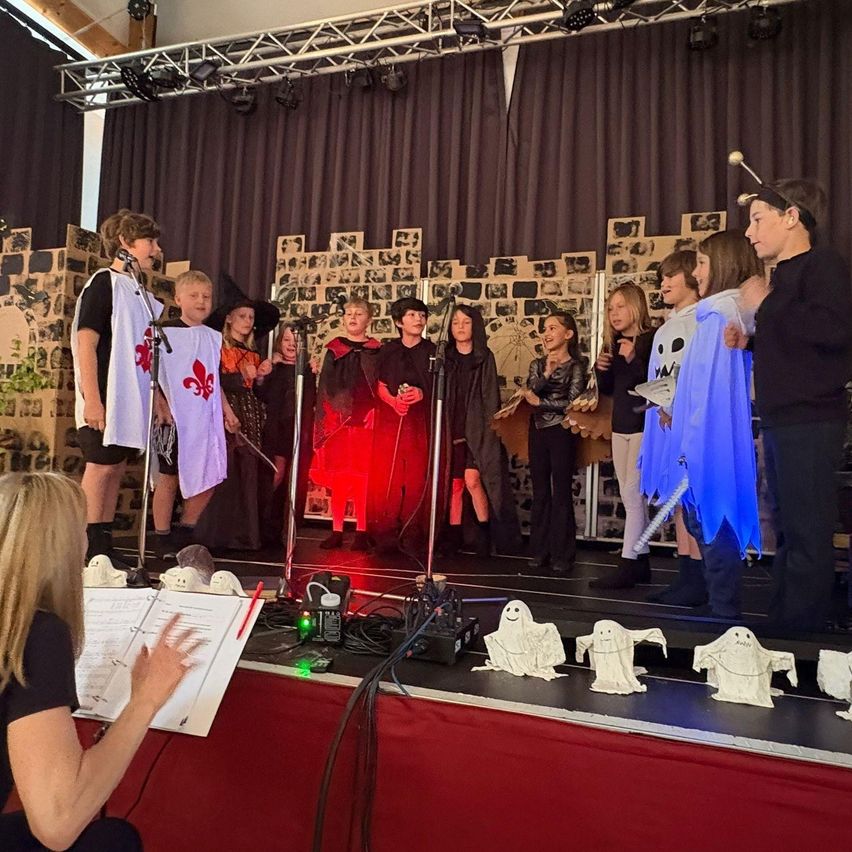 Eine Gruppe von Kindern führt ein Theaterstück auf der Bühne auf, gekleidet in Kostüme. Eine Frau beobachtet von der Seite mit einem Musikbuch. Rote Beleuchtung und Requisiten sind sichtbar.