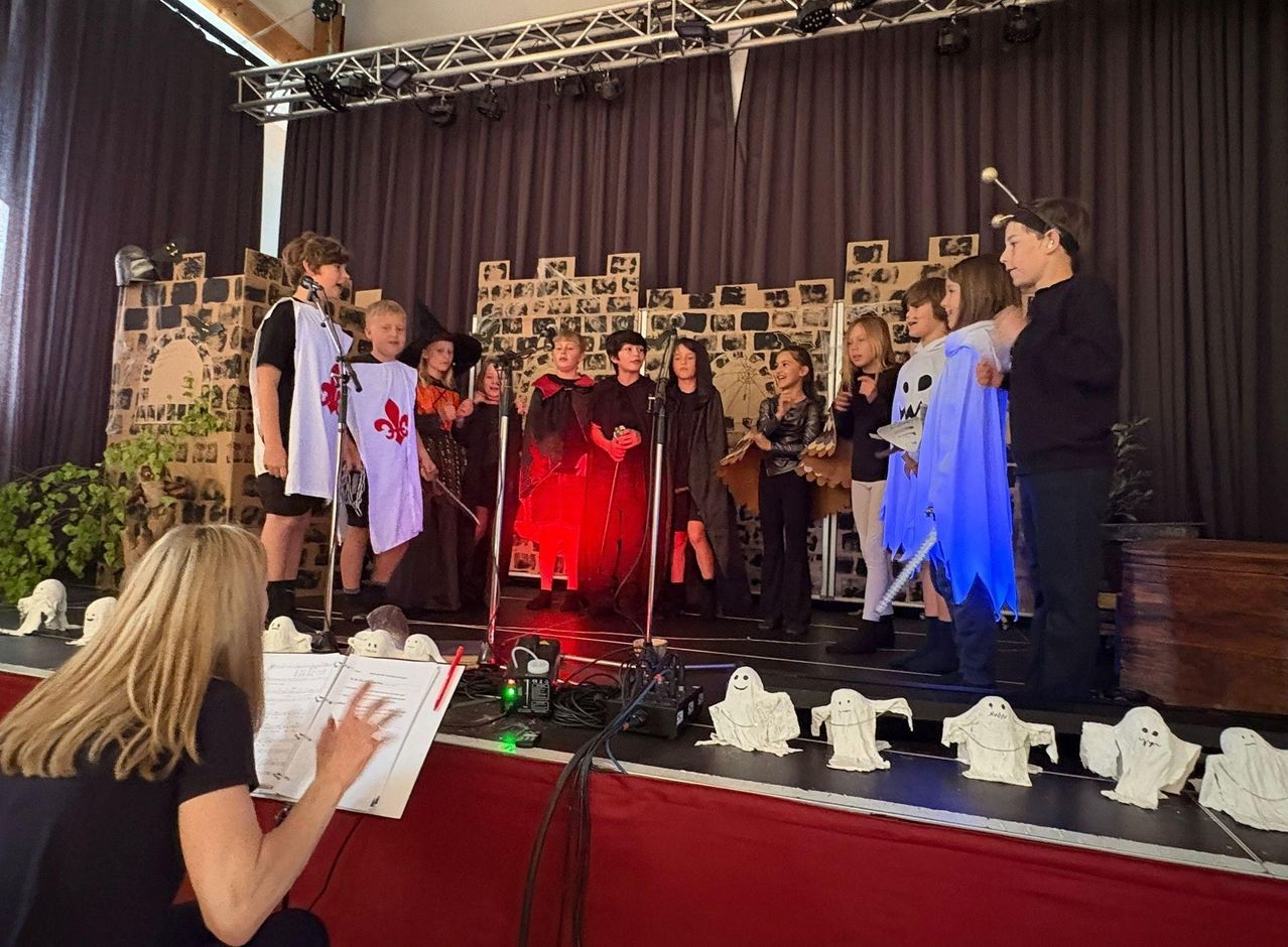 Eine Gruppe von Kindern führt ein Theaterstück auf der Bühne auf, gekleidet in Kostüme. Eine Frau beobachtet von der Seite mit einem Musikbuch. Rote Beleuchtung und Requisiten sind sichtbar.