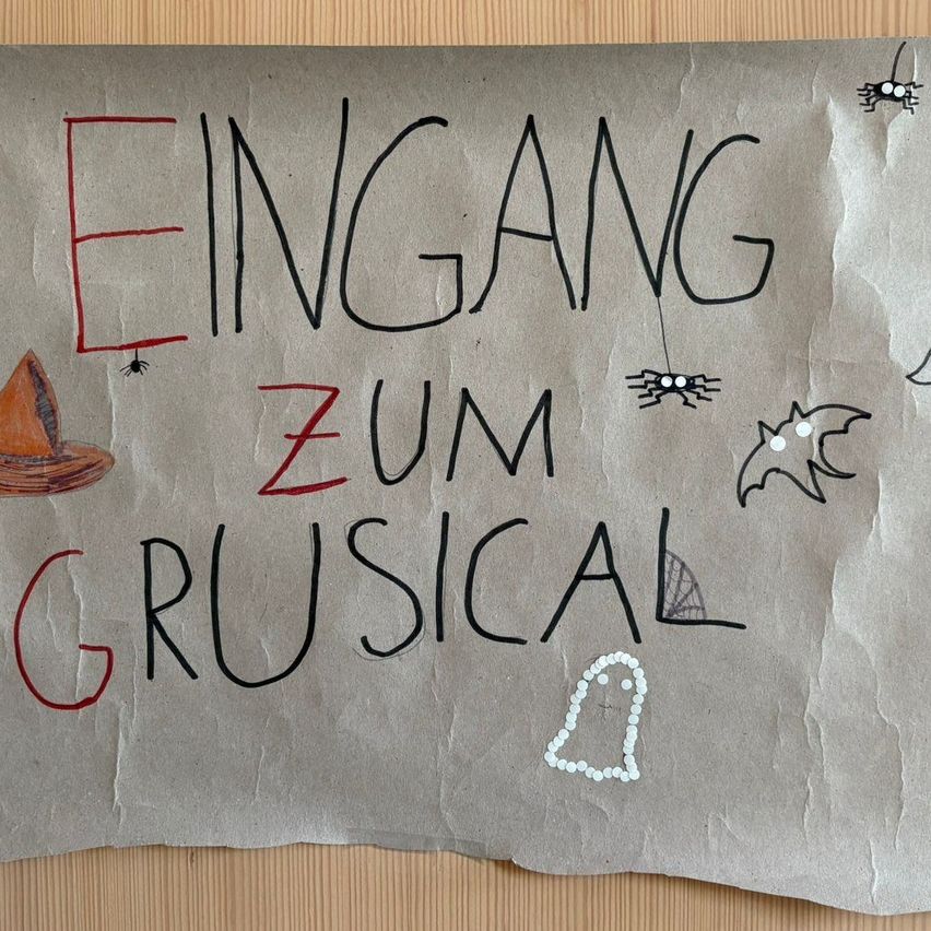 Ein Papierschild mit den Worten 'Eingang zum Grusical' in Schwarz und Rot geschrieben, mit Halloween-Dekorationen wie einer Hexenmütze und Fledermäusen.