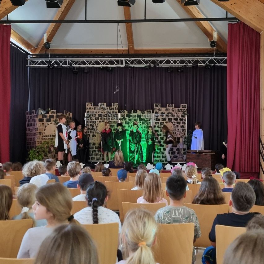 Eine Schulaufführung mit Kindern auf der Bühne, einem grünen Hintergrund und einem zuschauenden Publikum. Die Bühne hat eine Ziegelwand und eine Person in Grün.