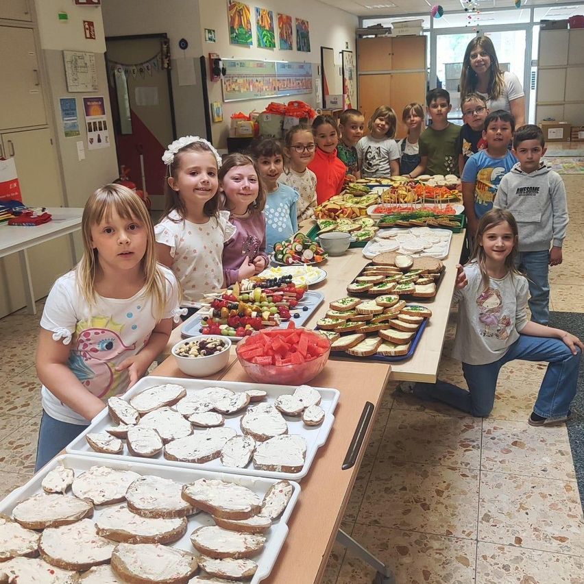Eine Gruppe von Kindern in einem Klassenzimmer posiert für ein Foto an einem Tisch, der mit verschiedenen Lebensmitteln gefüllt ist, einschließlich Sandwiches, Früchten und Keksen.
