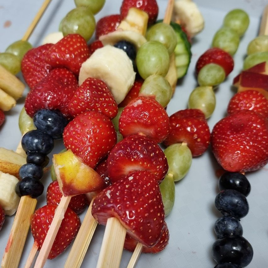 Obstspieße mit Erdbeeren, Trauben, Blaubeeren und Bananen auf Stöcken. Eine Vielfalt an Früchten auf einer weißen Oberfläche.