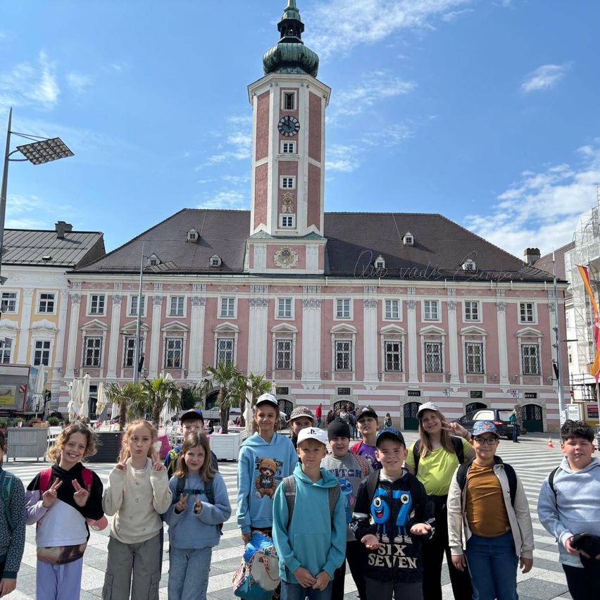 Eine Gruppe von Kindern steht vor einem großen Gebäude mit einem Turm. Das Gebäude hat viele Fenster und ist rosa gestrichen. Die Kinder tragen Mützen und Rucksäcke.
