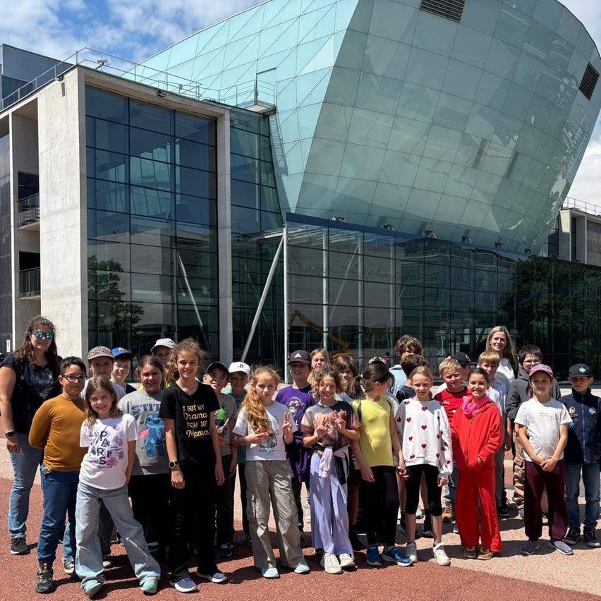 Eine Gruppe von Kindern und Erwachsenen posiert für ein Foto vor einem modernen, glasverkleideten Gebäude an einem sonnigen Tag.