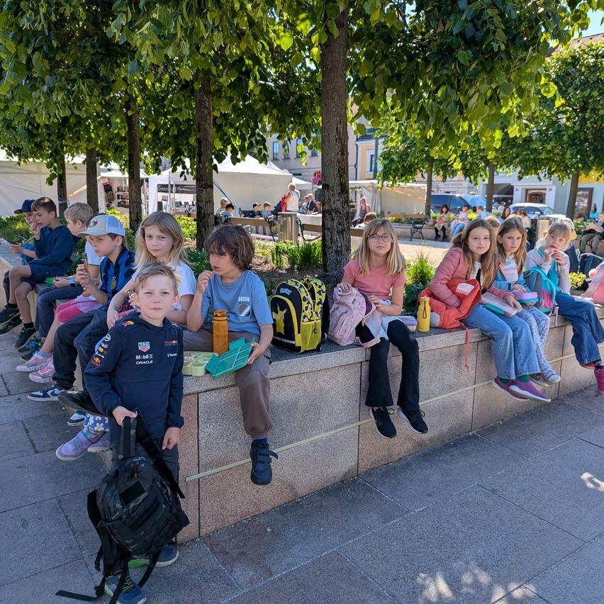 Eine Gruppe von Kindern sitzt auf einer Mauer in einem Park mit Bäumen im Hintergrund. Einige essen Snacks und tragen Rucksäcke.