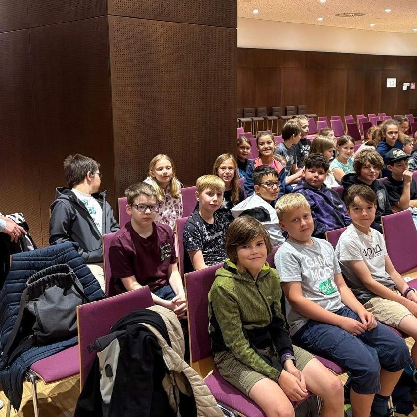 Ein Klassenzimmer mit Kindern in Reihen von lila Stühlen, einige tragen Brillen. Hinter ihnen ist eine Holzwand. Ein Junge trägt eine grüne Kapuzenjacke.