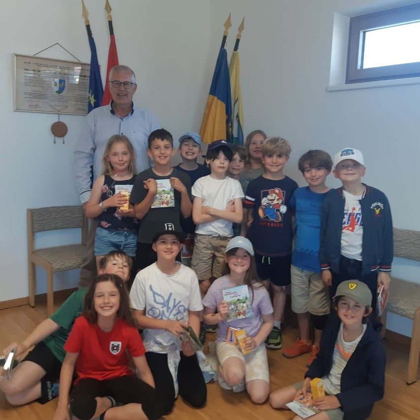 Eine Gruppe von Kindern, begleitet von einem Erwachsenen, posiert für ein Foto, während sie Bücher halten. Sie stehen und sitzen vor Flaggen und einem Fenster.