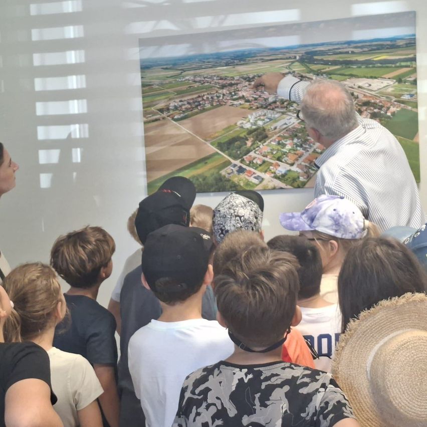 Eine Gruppe von Menschen betrachtet eine Luftaufnahme eines Dorfes. Ein älterer Mann zeigt auf das Bild an der Wand.