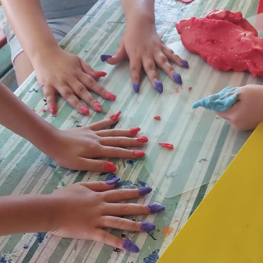 Kinderhände sind mit roter und lila Farbe auf einem mit einem gestreiften Tuch bedeckten Tisch bemalt. Ein Kind hält ein blaues Objekt.