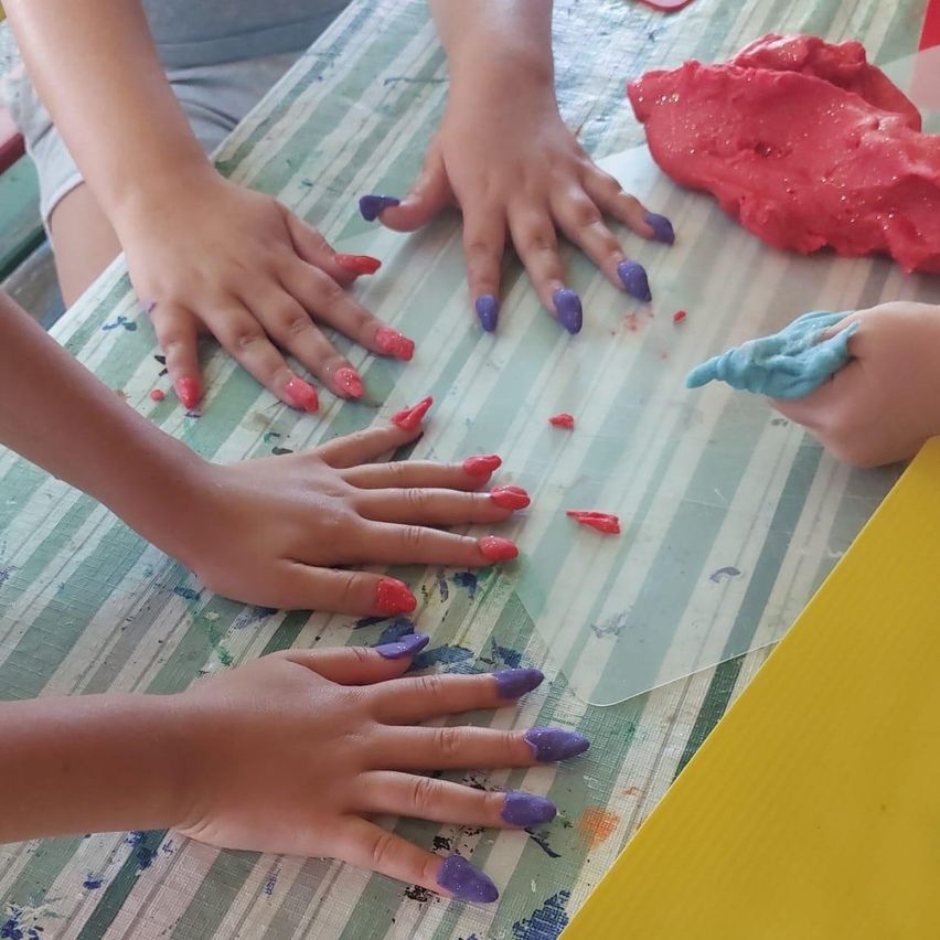 Kinderhände sind mit roter und lila Farbe auf einem mit einem gestreiften Tuch bedeckten Tisch bemalt. Ein Kind hält ein blaues Objekt.