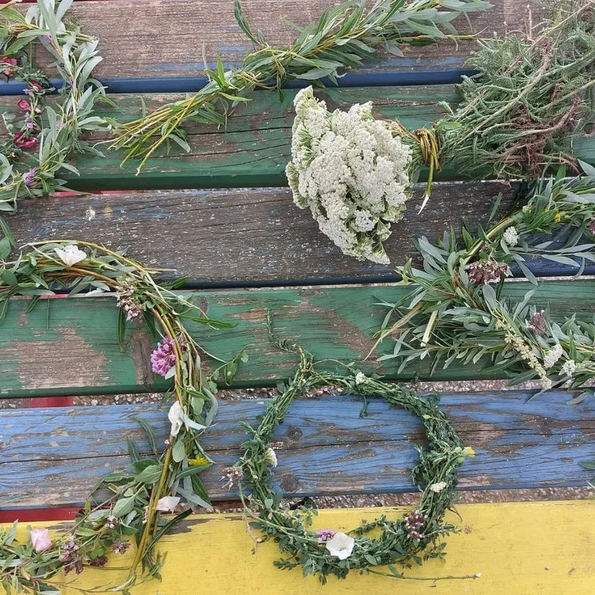 Mehrere Kränze aus verschiedenen grünen Pflanzen und Blumen sind auf einer hölzernen Bank angeordnet. Die Bank hat eine mehrfarbige Lackierung.