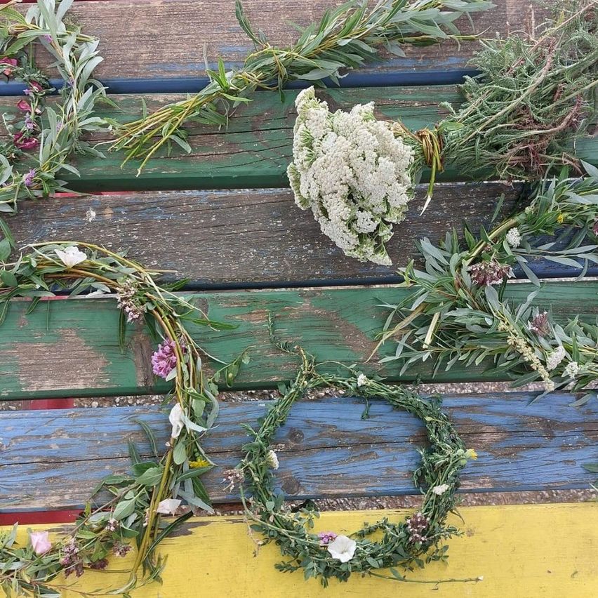 Mehrere Kränze aus verschiedenen grünen Pflanzen und Blumen sind auf einer hölzernen Bank angeordnet. Die Bank hat eine mehrfarbige Lackierung.