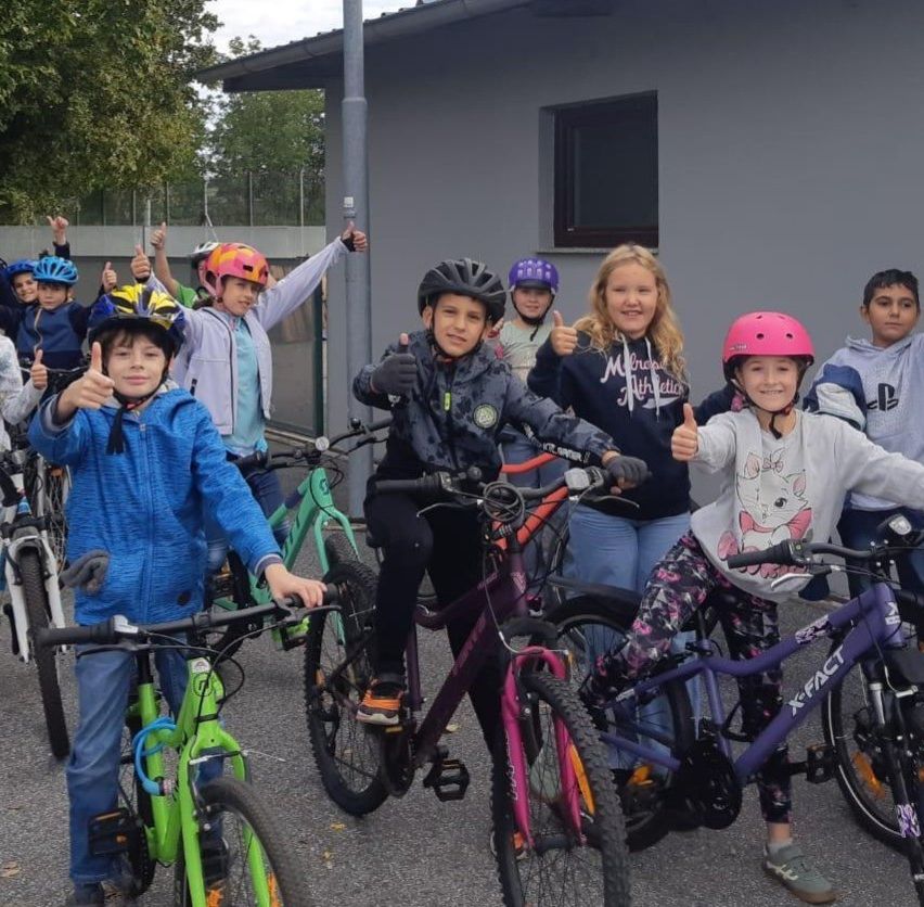 Eine Gruppe von Kindern, alle mit Helmen, posiert für ein Foto mit ihren Fahrrädern. Sie stehen auf einem Parkplatz.
