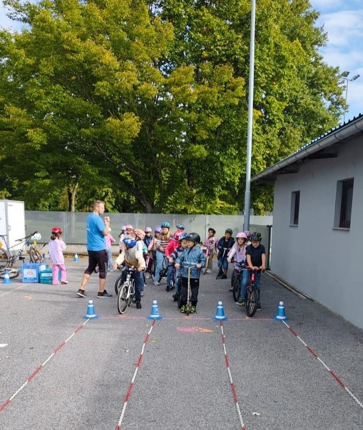 Eine Gruppe von Kindern mit Helmen fährt auf Fahrrädern in einem Parkplatz, begleitet von einem Mann in einem blauen T-Shirt. Bäume und ein Gebäude sind im Hintergrund zu sehen.