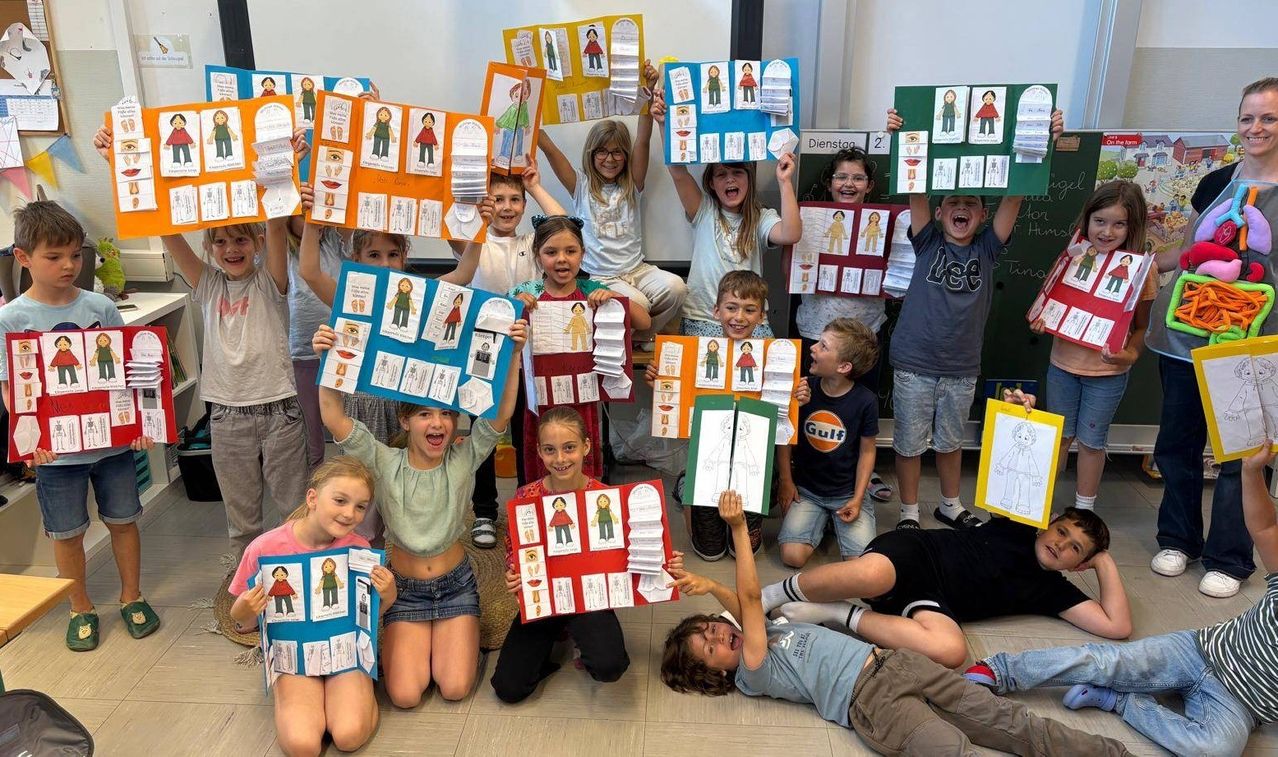 Eine Gruppe von Kindern posiert für ein Foto mit bunten pädagogischen Postern über den menschlichen Körper. Sie halten die Poster mit Illustrationen von Körperteilen hoch.