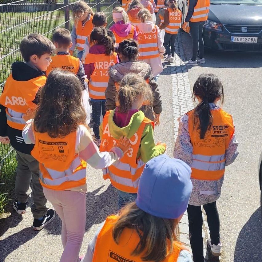 Eine Gruppe von Kindern in orangefarbenen Warnwesten geht auf einem Bürgersteig. Ein Auto ist hinter ihnen auf der Straße geparkt.