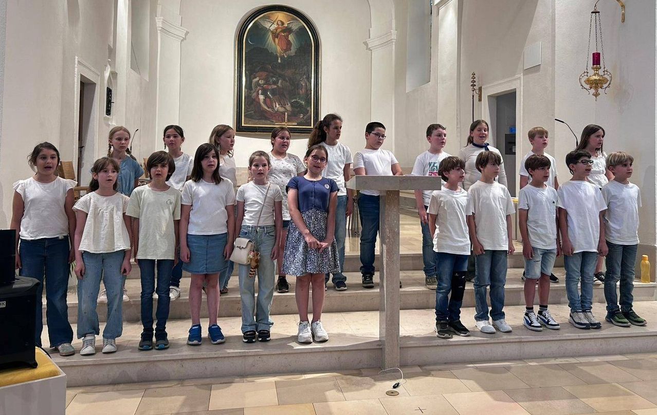 Eine Gruppe von Kindern in weißen Hemden steht in einer Kirche, möglicherweise singend, mit einer gemalten Wandmalerei dahinter.