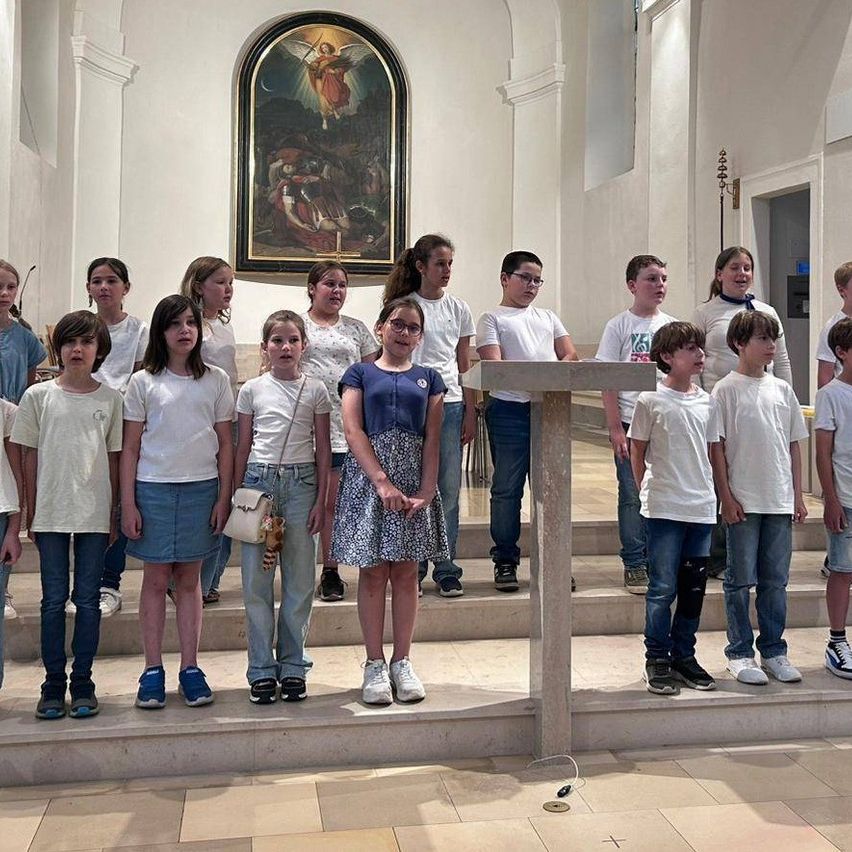 Eine Gruppe von Kindern in weißen Hemden steht in einer Kirche, möglicherweise singend, mit einer gemalten Wandmalerei dahinter.