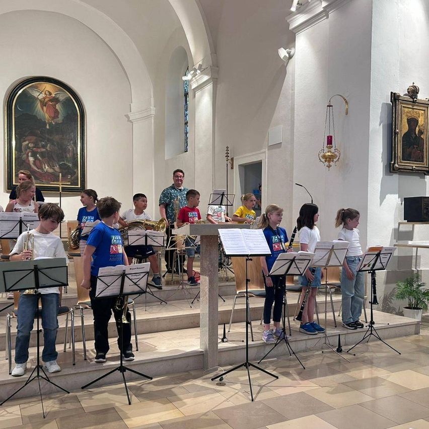 Eine Gruppe von Kindern, einige stehend und einige sitzend, spielen Musikinstrumente in einer Kirche. Sie schauen zum Altar, über dem ein Gemälde hängt.