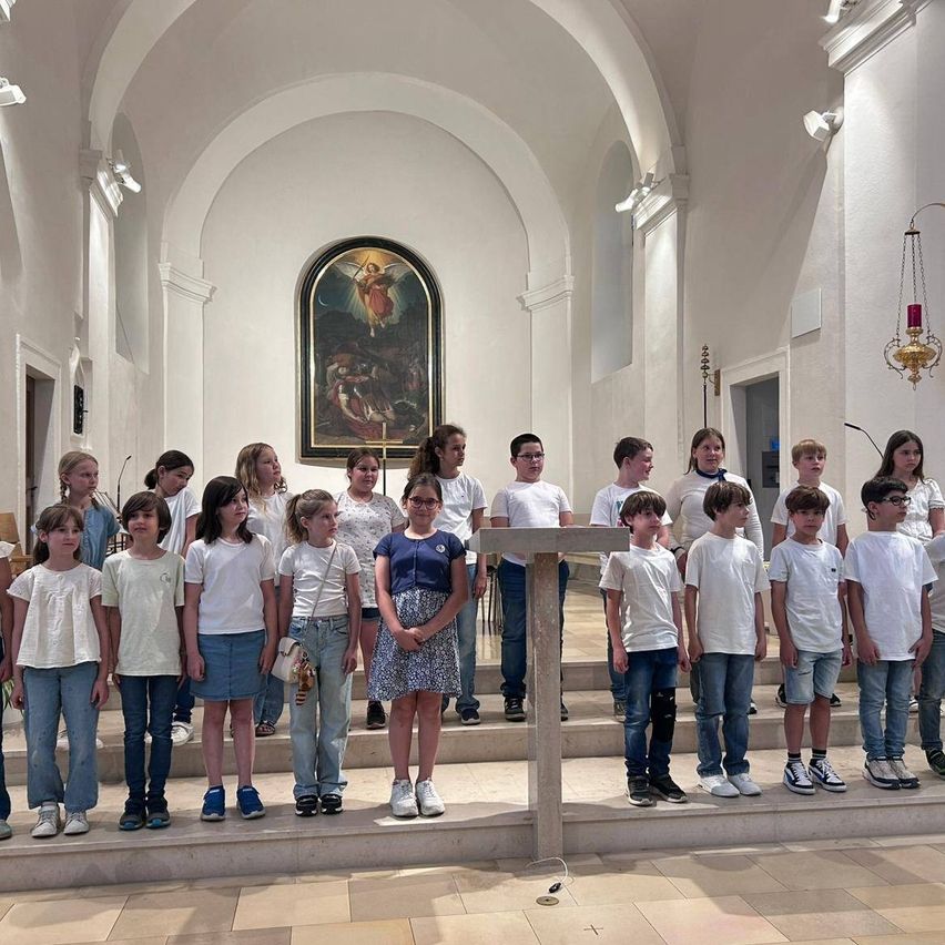 Ein Kinderchor steht auf den Stufen einer Kirche, gekleidet in Weiß, mit einem Kreuz davor und einem religiösen Gemälde dahinter.