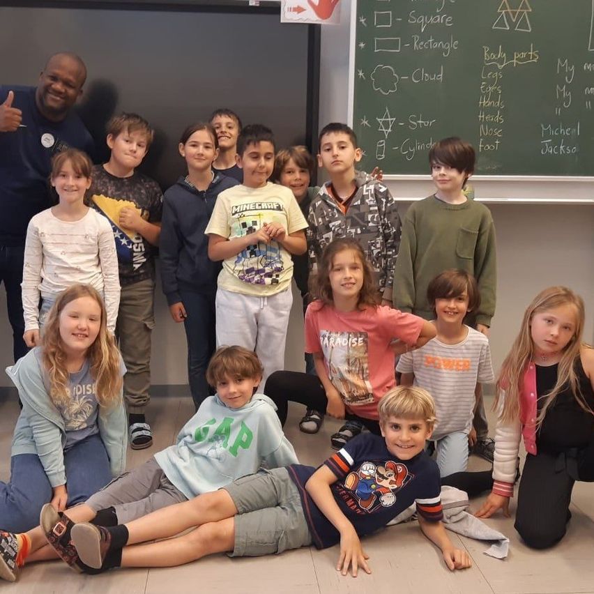Eine Gruppe von Kindern und einem Mann posiert für ein Foto im Klassenzimmer. Die Kinder sitzen und knien auf dem Boden. Der Mann lächelt und gibt einen Daumen hoch. Hinter ihnen steht eine Tafel mit einigen darauf geschriebenen Worten.