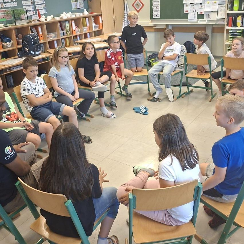 Ein Klassenzimmer mit Kindern in Stühlen im Kreis. Einige tragen Brillen, Turnschuhe und Armbänder. Ein Junge steht hinter ihnen, und Bücherregale sind in der Nähe.