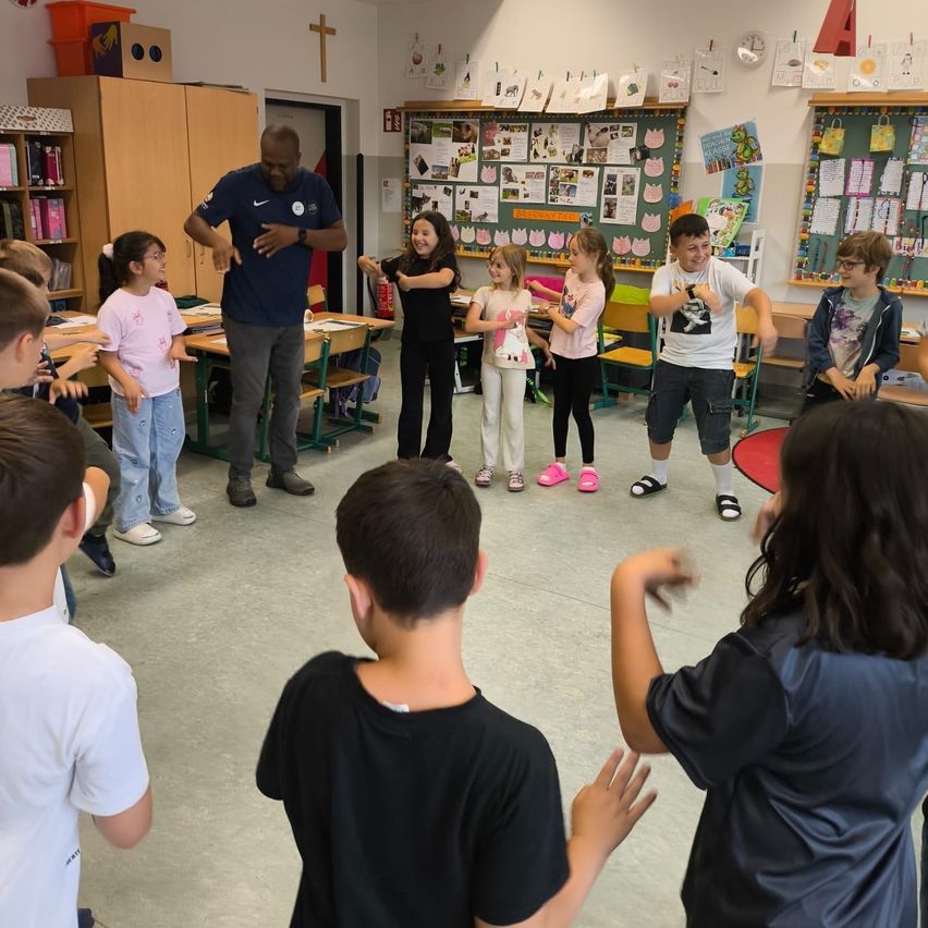 In einem Klassenzimmer machen eine Gruppe von Kindern und einem Mann eine Aktivität. Die Kinder stehen im Kreis, während der Mann sie anführt. Die Kinder tragen Freizeitkleidung. Das Klassenzimmer hat Schränke, Regale und eine Bulletin-Board.