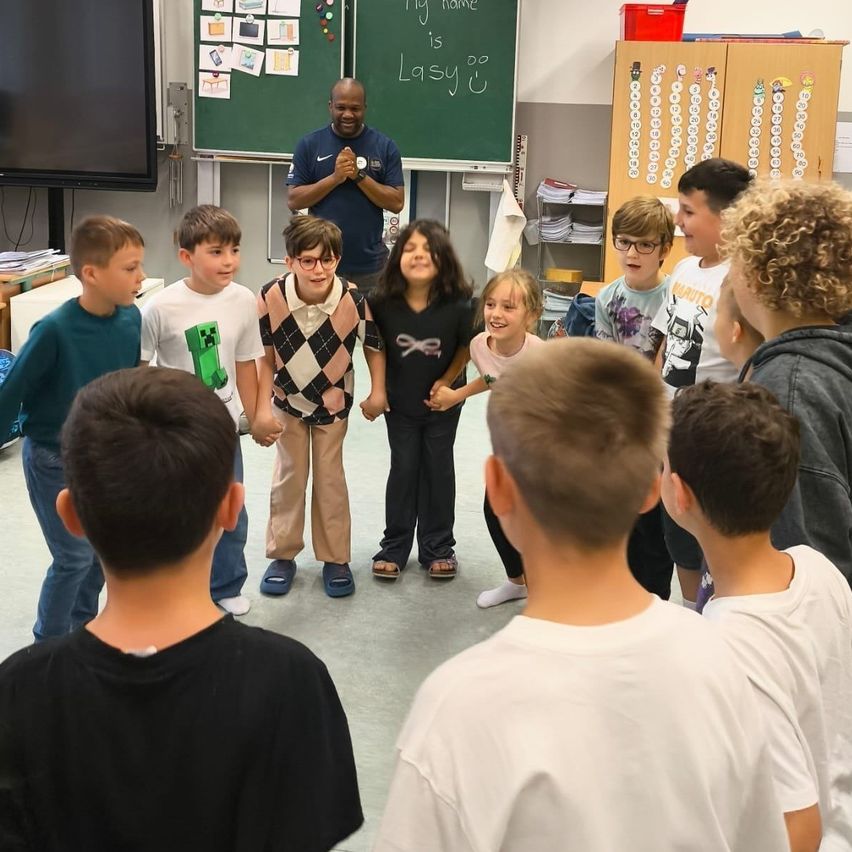 Schüler stehen in einem Klassenzimmer im Kreis mit einem Lehrer, eine Tafel hinter ihnen. Der Lehrer klatscht.