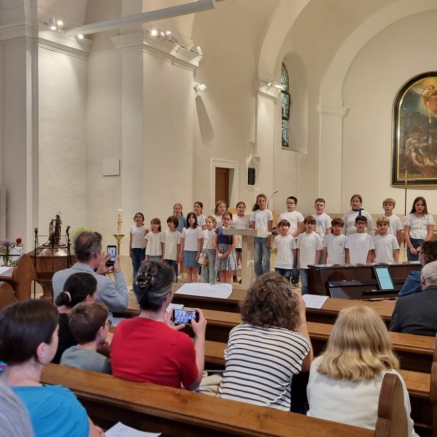 Ein Chor von Kindern in weißen Hemden und Röcken oder Shorts steht in einer Kirche, einige Erwachsene sitzen und andere halten Kameras. Ein Klavier steht dahinter, und eine Statue und ein Gemälde sind an der Wand.