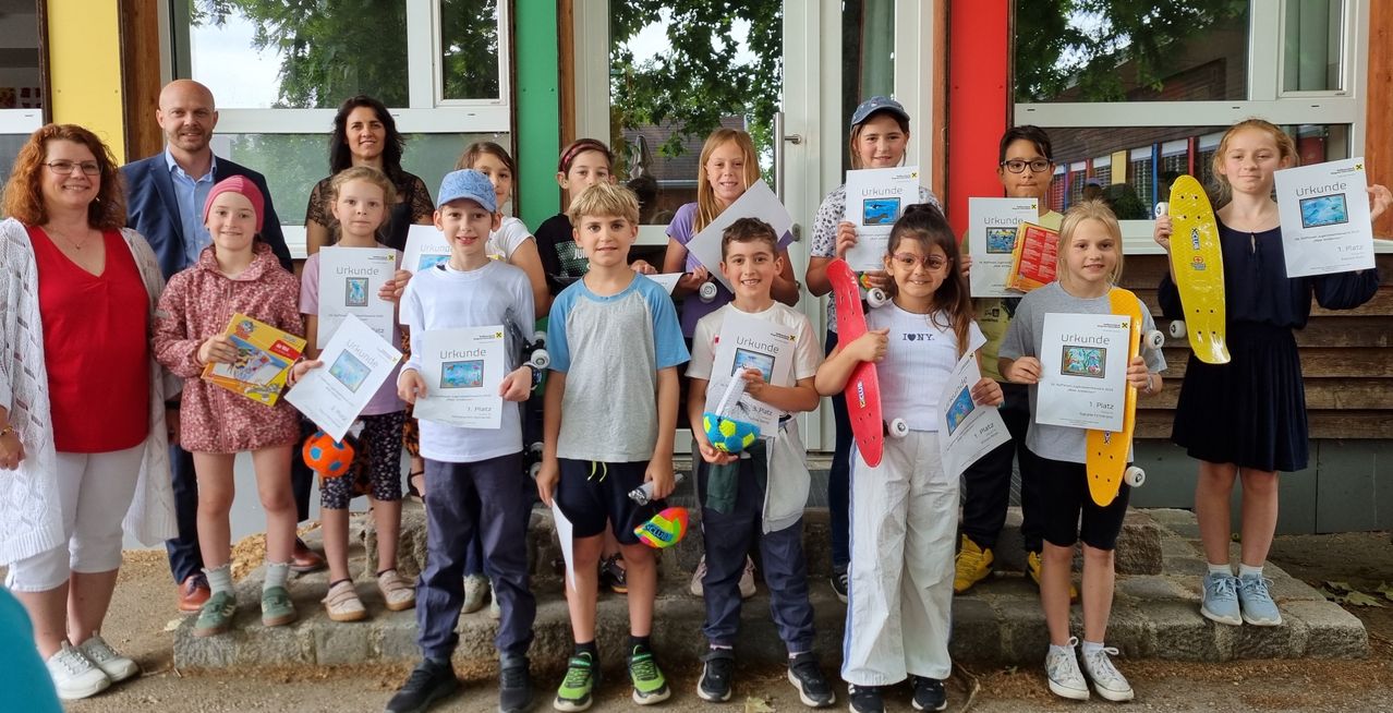 Eine Gruppe junger Kinder, lässig gekleidet, steht draußen und hält Urkunden. Einige tragen Hüte und Brillen. Sie lächeln und posieren für ein Foto. Dahinter halten Erwachsene Papiere.