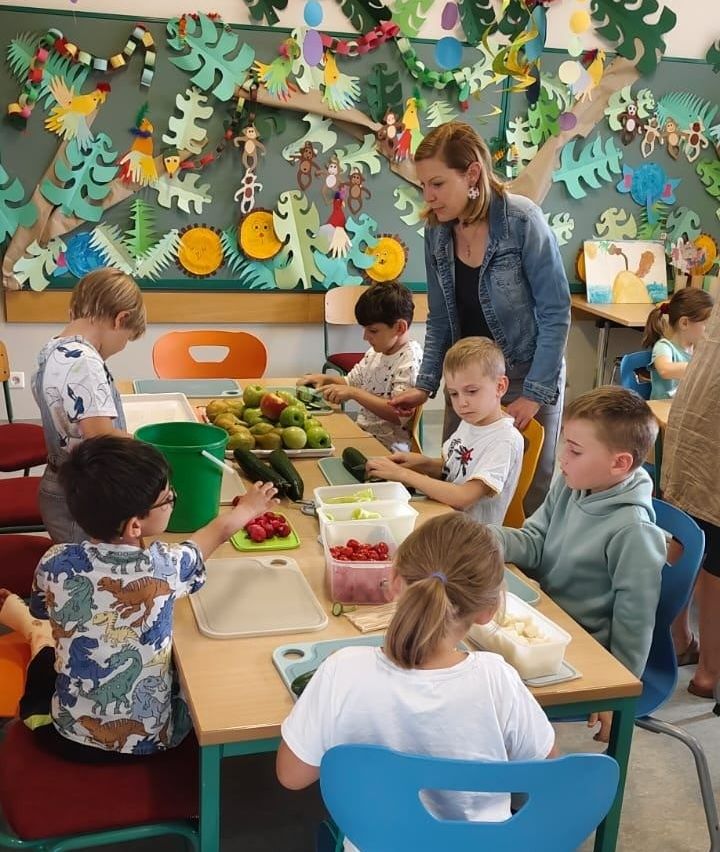 Eine Gruppe von Kindern in einem Klassenzimmer schneidet Obst und Gemüse, während eine Lehrerin sie beaufsichtigt. Der Raum hat eine dschungelthematische Wanddekoration.