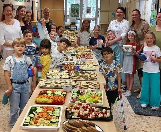 Eine Gruppe von Kindern und Erwachsenen steht hinter einem langen Tisch voller Essen und lächelt. Der Tisch hat Sandwiches, Salate und andere Lebensmittel.