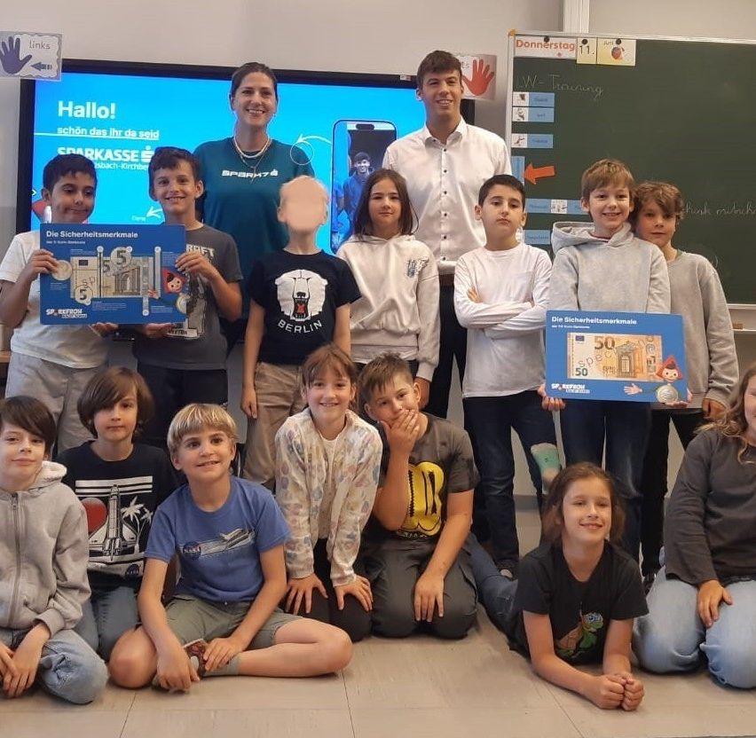 Eine Gruppe von Schülern im Klassenzimmer, lächelnd und posierend für ein Foto. Einige halten blaue Boxen mit einer 50-Euro-Note, andere ein Buch. Zwei Erwachsene stehen dahinter. Ein Tafel mit einem roten Pfeil und anderen Gegenständen ist sichtbar.