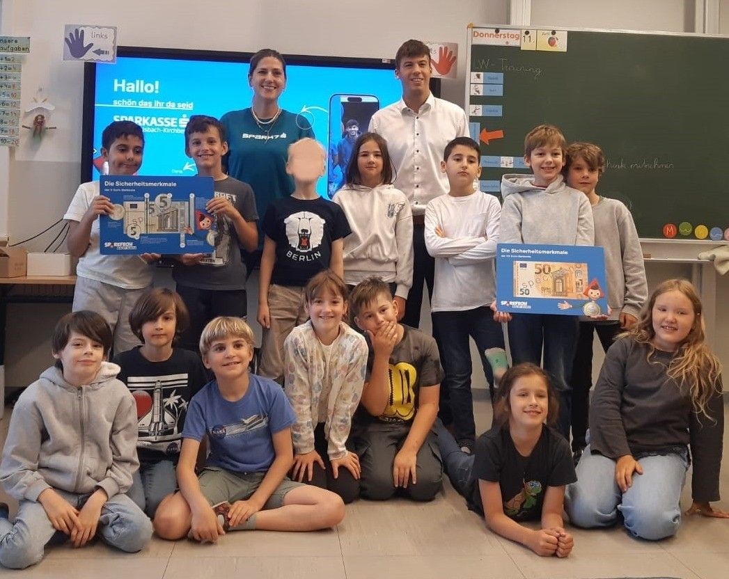 Eine Gruppe von Schülern im Klassenzimmer, lächelnd und posierend für ein Foto. Einige halten blaue Boxen mit einer 50-Euro-Note, andere ein Buch. Zwei Erwachsene stehen dahinter. Ein Tafel mit einem roten Pfeil und anderen Gegenständen ist sichtbar.