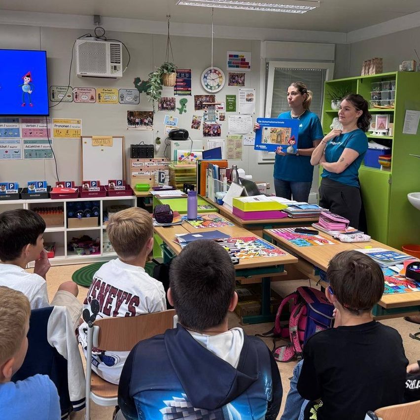 Zwei Frauen stehen in einem Klassenzimmer und halten ein großes blaues Plakat in der Hand, während eine Gruppe von Jungen aufmerksam zuhört. Hinter ihnen schmücken Regale mit Büchern und eine Uhr die Wand, und ein Fernsehbildschirm zeigt ein Bild.