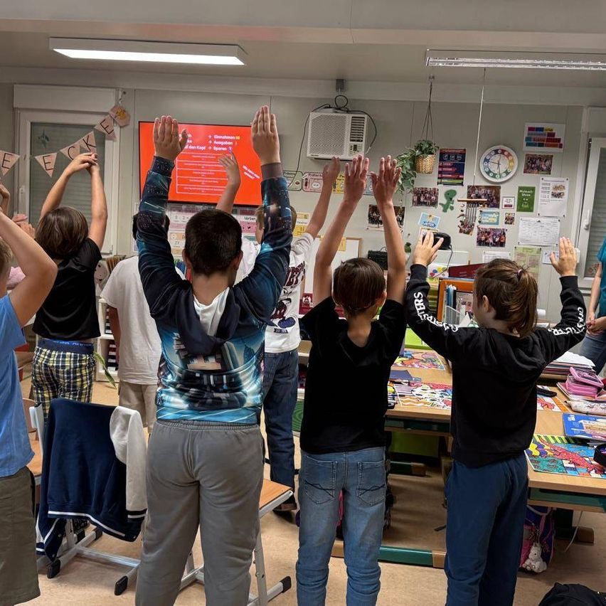 Eine Gruppe von Schülern im Klassenzimmer mit erhobenen Händen, die auf einen Bildschirm schauen. Sie tragen Freizeitkleidung, und ein bunter Banner ist im Hintergrund.