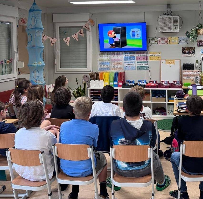 In einem Klassenzimmer sitzen eine Gruppe von Kindern auf Stühlen vor einem montierten Fernseher, der einen Cartoon-Charakter zeigt. Sie scheinen aufmerksam zu schauen, mit verschiedenen Lehrmaterialien und Dekorationen an den Wänden.