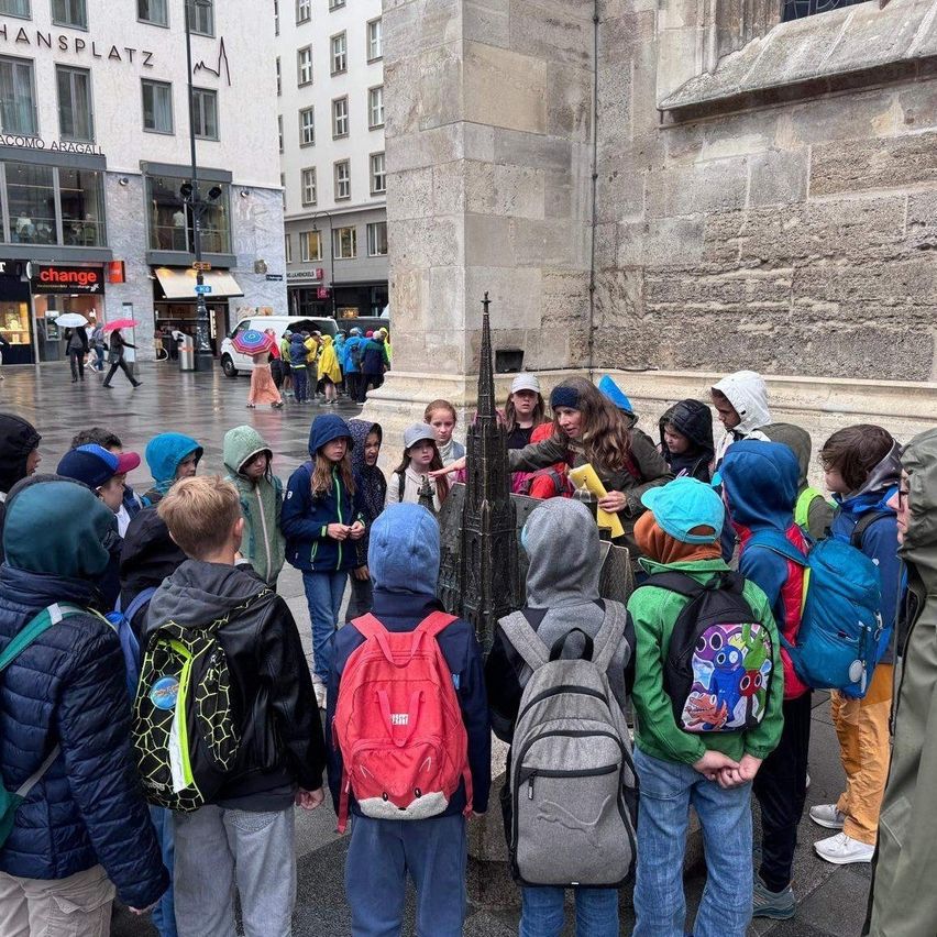 Eine Gruppe von Kindern in Regenkleidung und Rucksäcken versammelt sich um ein Steinmonument. Sie befinden sich auf einer nassen Straße mit einer Menge von Menschen und Gebäuden im Hintergrund.