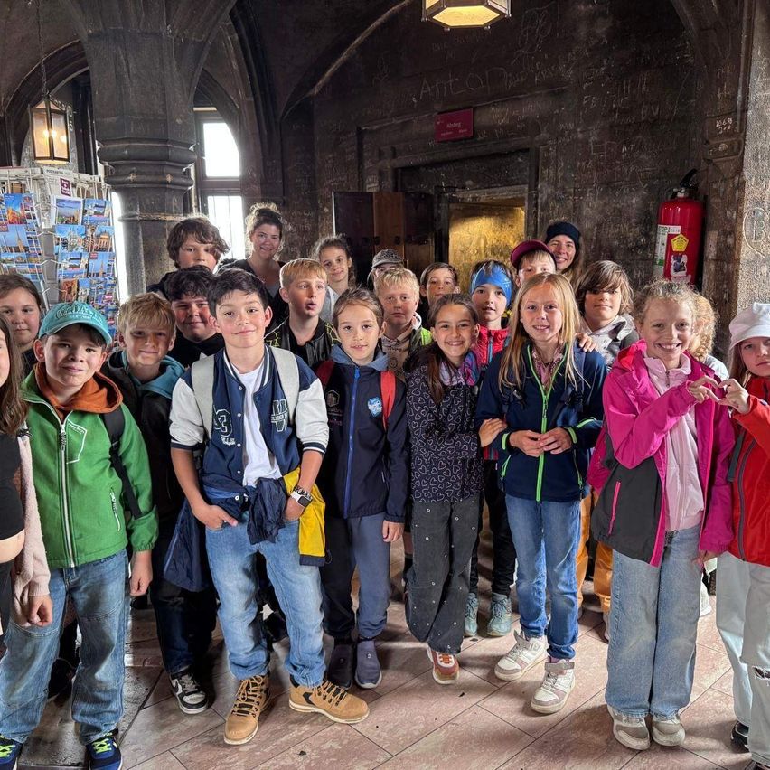Eine Gruppe von Kindern, einige mit Mützen, lächeln und posieren für ein Foto in einem Raum mit gefliesten Böden und Bogenfenstern.