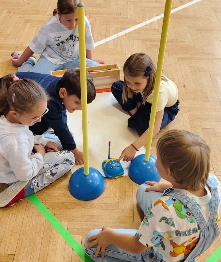 Eine Gruppe von Kindern spielt mit einem Spielzeugauto und Luftballonpfosten auf einem Holzboden. Ein Kind fährt das Spielzeugauto, während die anderen zuschauen.