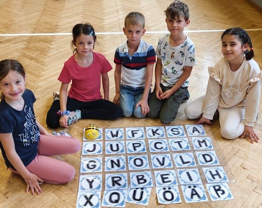 Fünf Kinder sitzen um ein Wortspiel auf einem Holzboden und lächeln. Buchstaben von W bis B sind auf dem Boden angezeigt. Ein Bienenspielzeug ist links platziert.
