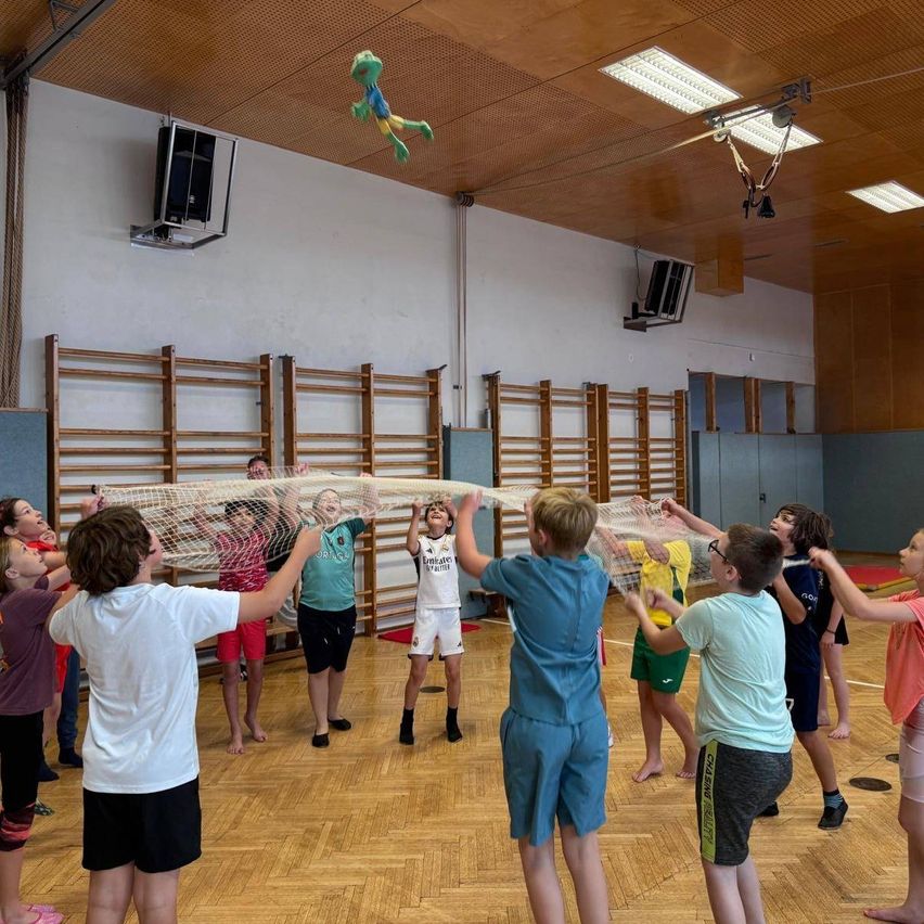 Eine Gruppe von Kindern in einer Turnhalle, die mit einem großen Netz spielen, einige tragen Sportuniformen. Im Hintergrund ist ein grüner Frosch von der Decke aufgehängt.