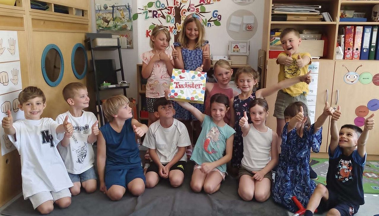Gruppe von Kindern in einem Klassenzimmer, die eine Twister-Spielplatte hochhalten, mit einer lächelnden und fröhlichen Atmosphäre.