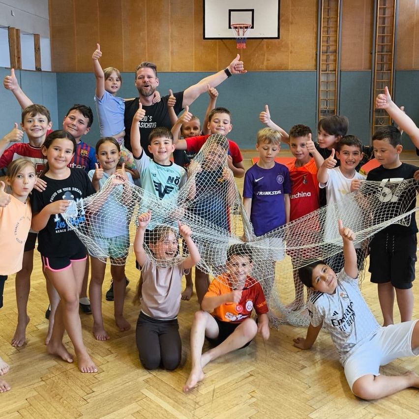 Eine Gruppe von Kindern, Jungen und Mädchen, lächelt und posiert für ein Foto in einer Turnhalle. Ein Mann hält eine große Netz in der Mitte.