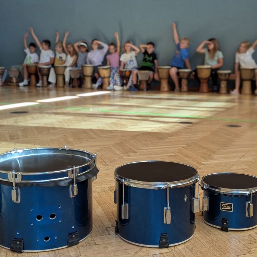 Eine Gruppe von Kindern sitzt mit erhobenen Armen auf Trommeln, drei blaue Trommeln im Vordergrund.